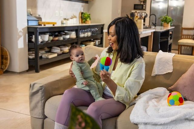 Woman holding baby on sofa shaking colorful rattle for infant bonding and play
