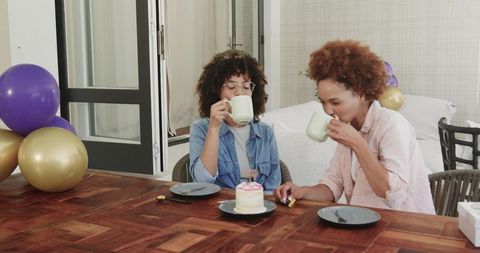 Two women sharing coffee and single-tier celebration cake in sunlit sunroom with balloons