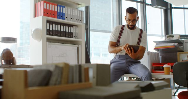 Focused young biracial man reviews a tablet in an office. He's surrounded by shelves and documents, indicating a busy work environment.
