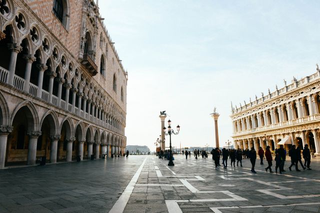 Tourists exploring the historic St. Mark's Square in Venice under a clear sky. The scene includes iconic Venetian architecture, lampposts, and a clear sky, making it perfect for travel advertisements, blogs, tourism brochures, cultural studies, and guides.