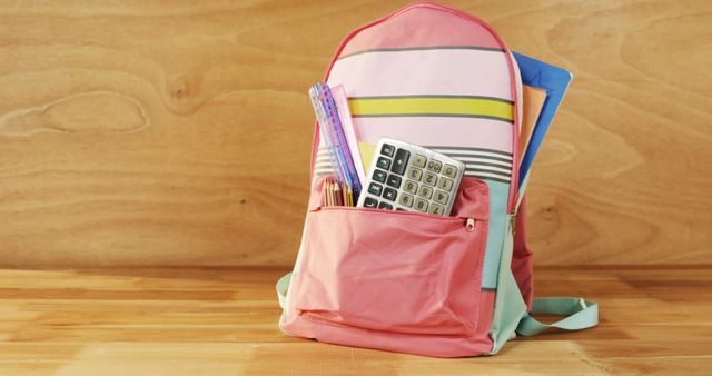 A vibrant pink backpack is sitting on a wooden desk. The backpack has various school supplies, including a calculator, colorful rulers, and notebooks peeking out of its pockets. This image is ideal for back-to-school promotions, educational content, student life features, and advertisements for school supplies and stationery.