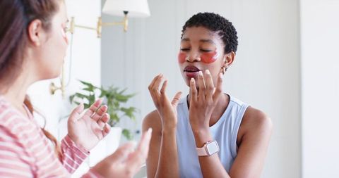 Diverse friends pampering with under-eye gel patches, chatting and checking smartwatch