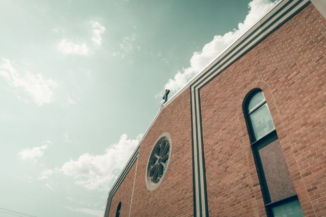 This image captures the exterior of a church with brick walls, featuring a prominent rose window and a cross on the roof. The sky above is filled with scattered clouds. This image can be used for articles on religious architecture, spirituality, or community gatherings. It can also serve marketing purposes for church websites and architectural designs.