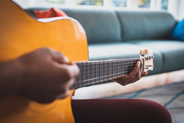 African American woman sitting on a couch playing an acoustic guitar, focusing on her hands and the guitar strings. Ideal for use in articles or advertisements about music, hobbies, relaxation, home activities, quarantine, and lifestyle.