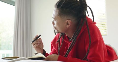 African american man lying on bed journaling and planning in bright bedroom, balcony view