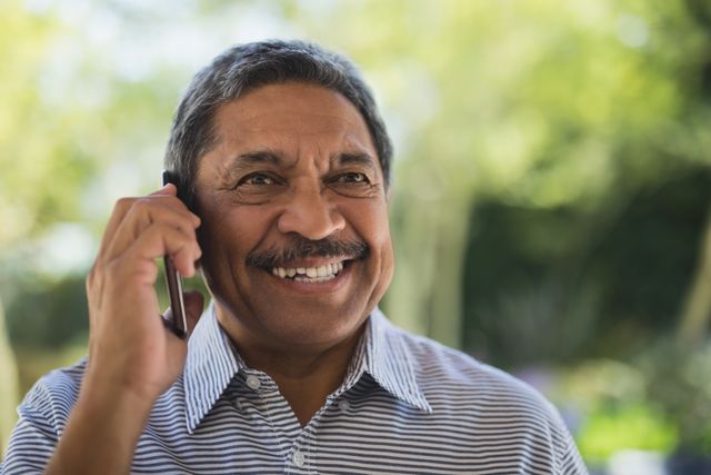 Senior man smiling while talking on mobile phone outdoors. Ideal for use in advertisements, articles, and promotions related to senior lifestyle, communication technology, and happiness in older age. Suitable for illustrating concepts of staying connected, enjoying life, and modern technology use among elderly individuals.