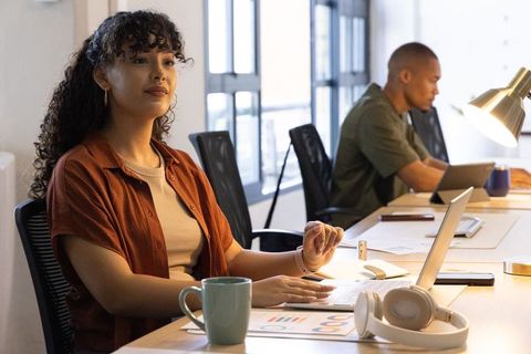 Young woman working at shared coworking desk with laptop, charts, coffee, daylight