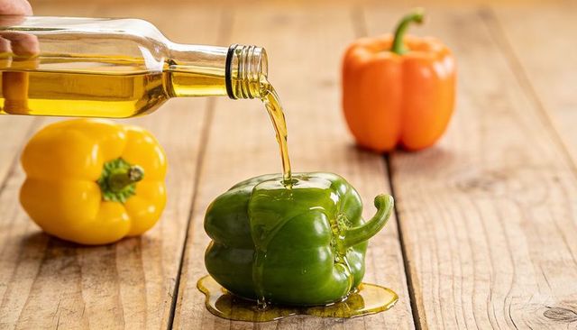 Olive oil pouring over green bell pepper with yellow and orange peppers on wooden plank