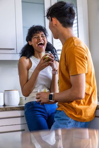 Laughing african american woman and asian man holding drinks in bright kitchen lifestyle