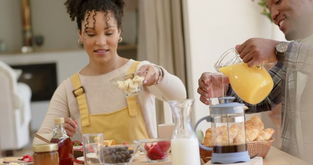 Couple enjoying a wholesome breakfast together at home. Fresh ingredients on the table, including orange juice, a French press, croissants, and fruit. Ideal for use in advertisements or articles about healthy eating, family life, or domestic bliss.