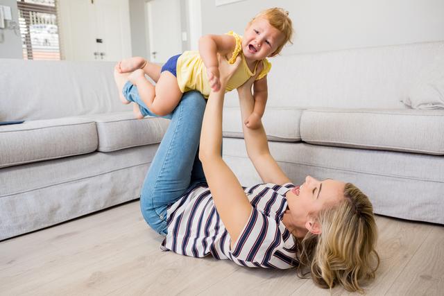 Mother lifting baby girl in air while lying on floor in living room. Ideal for use in parenting blogs, family lifestyle articles, advertisements for home products, or promotional materials for family-oriented services.