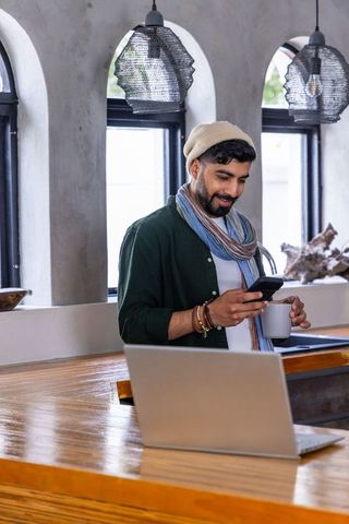 Asian man using smartphone and laptop in minimalist cafe holding ceramic mug in warm natural light
