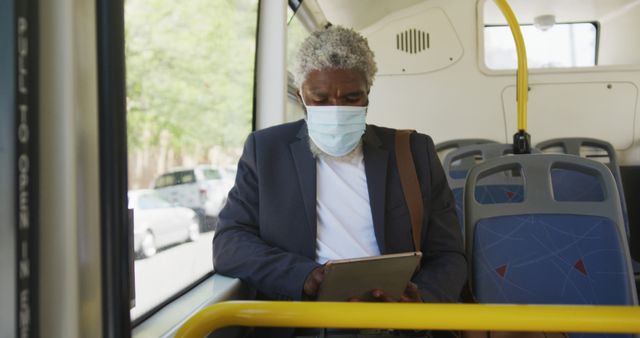 Elderly man in formal attire wearing a mask, sitting on a bus and using a digital tablet. Image can be used for promoting public transport safety protocols, technology use among seniors, or pandemic-related commuting practices.