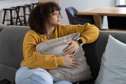Contemplative woman hugging textured beige cushion on sofa in cozy mustard sweater