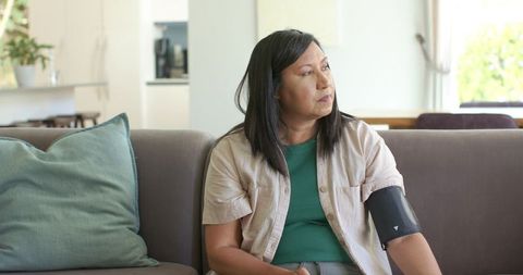 Asian mature woman sitting on sofa monitoring blood pressure with home cuff, looking thoughtful