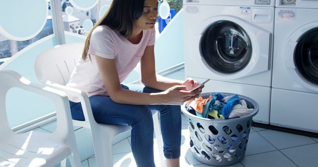 Happy biracial woman using smartphone sitting by laundry basket in sunny laundromat, copy space. Communication, laundry, washing machine, domestic chores and local business, unaltered.