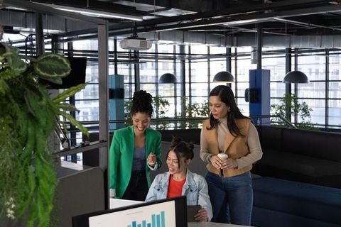 Three diverse women collaborating and analyzing data on monitor in modern open-plan office