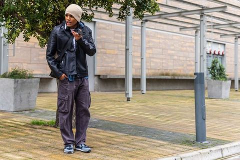 Man speaking into smartphone while standing on city plaza in beanie and leather jacket