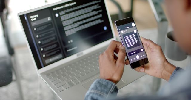 Man engaging with smartphone while using laptop in office. Ideal for illustrating modern workspaces, remote work concepts, digital communication, multitasking in business, and technology use in professional settings.