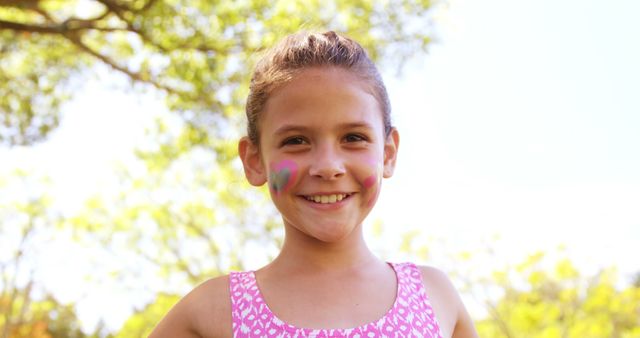 A young Caucasian girl with face paint smiles brightly in an outdoor setting, with copy space. Her cheerful expression and the face paint suggest she's enjoying a festive occasion or a playful day outside.