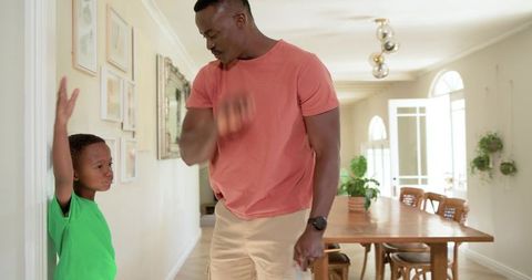 African american father guiding young son raising hand in hallway near dining table