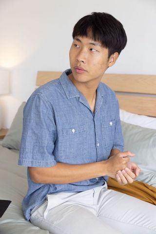 Contemplative asian man sitting on bed wearing blue shirt clasping hands looking left