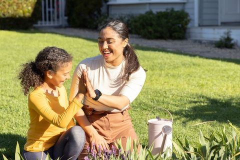 Asian mother and african american daughter laughing and high-fiving in sunlit garden