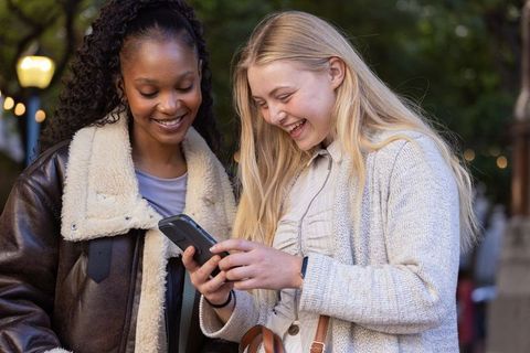 Multicultural friends sharing smartphone and laughing on city street at dusk