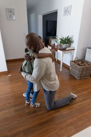African american father kneeling and hugging daughter with backpack in modern home