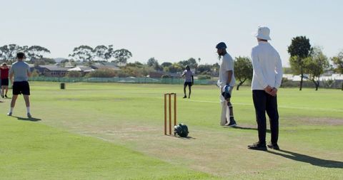 Weekend cricket match with batter, umpire, wicket and helmet on suburban grassy pitch