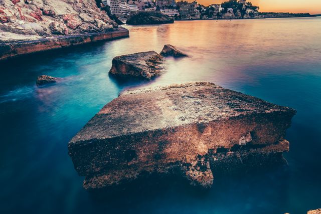 Calm seawater with submerged rocks bathed in the golden hues of the setting sun creates a serene coastal scene. This image is ideal for use in travel promotions, relaxation and meditation themes, or nature-focused advertisements.