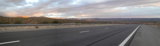 Long stretch of highway under dramatic sky with distant mountains and stunning sunset glow. Ideal for content related to road trips, travel, scenic views, and outdoor adventures. Useful for brochures, websites, and social media promoting road safety, exploring nature, or travel destinations.