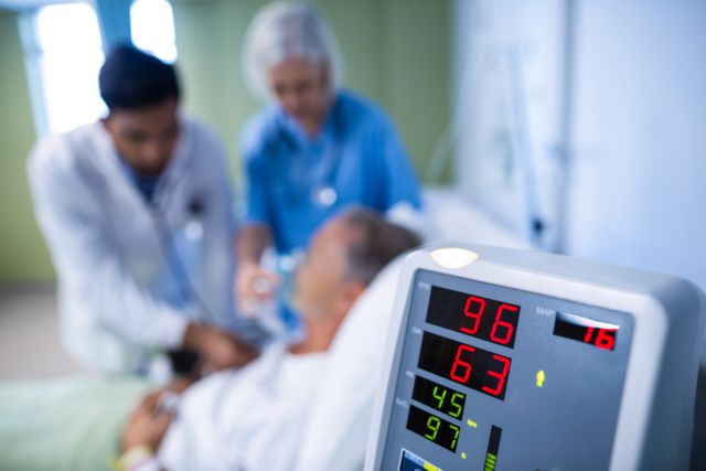 Heart rate monitor displaying vital signs in foreground while medical team treats patient in hospital bed. Useful for illustrating healthcare, emergency medical treatment, patient care, and hospital environments.