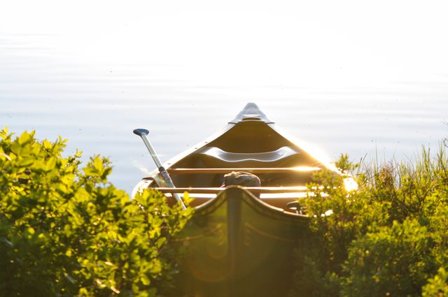 Empty canoe resting on rocky shore at sunrise, reflecting golden sunlight on calm water. Ideal for concepts of solitude, peace, and outdoor adventures. Perfect for promoting recreational activities, serene scenic views, and nature retreats.