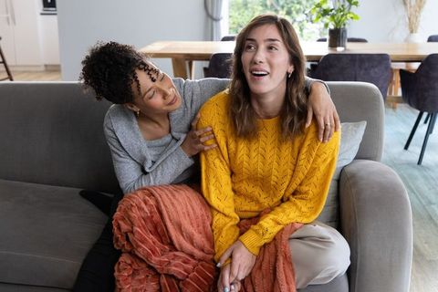 Multiracial friends laughing and cuddling on couch wearing cozy sweaters and rust blanket