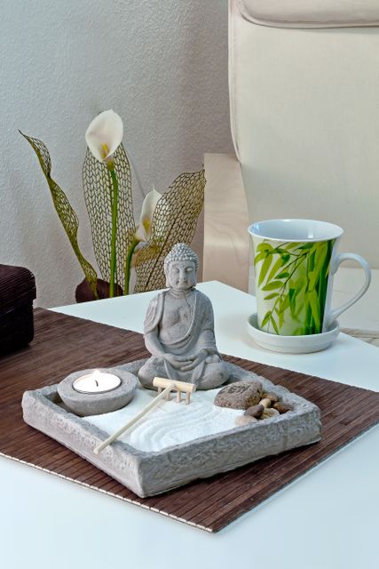 Peaceful Zen garden scene featuring a Buddha statue and a tea light, perfect for promoting relaxation and mindfulness. Set is complemented by a decorative teacup and elegant calla lilies in background. Ideal for use in wellness blogs, spa promotions, meditation resources, and home decor inspiration.