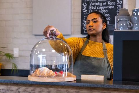 Indian barista placing glass cloche over croissant on rustic cafe counter, warm interior