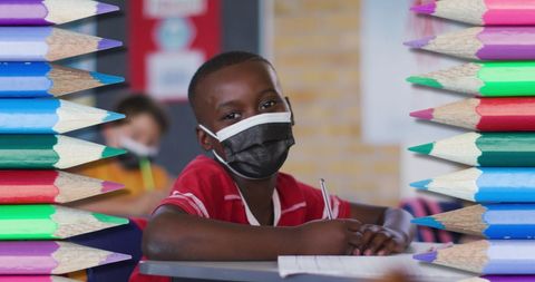 Student masked and focused, framed by colorful pencils