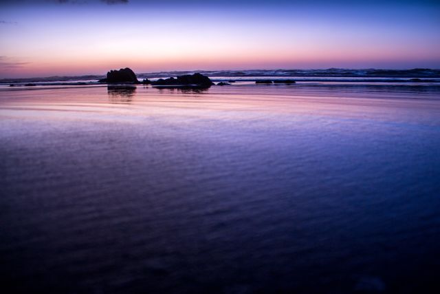This image showcases a serene ocean at sunset, with the sky displaying a gradient of colors from orange to deep blue. The calm water reflects these stunning colors. Rocks are silhouetted against the horizon, enhancing the tranquil ambiance. Ideal for backgrounds, travel blogs, meditation and relaxation themed projects, or advertisements promoting scenic destinations.