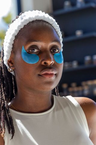 African woman wearing blue under-eye gel patches and headband preparing skincare routine