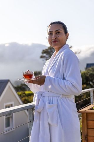 Asian woman enjoying morning tea on balcony in white bathrobe overlooking misty valley