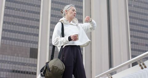 Mature commuter checking watch while holding coffee and duffel on city stairway