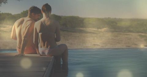 Couple sitting on infinity pool deck watching sunlit sandy shore during golden hour