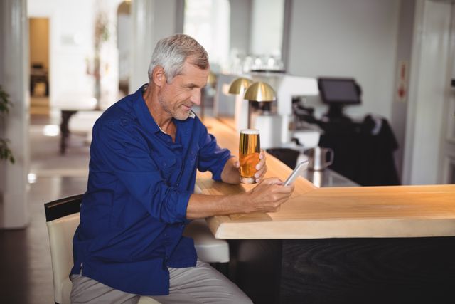 Mature man sitting at bar counter, using mobile phone while holding glass of beer. Ideal for themes related to leisure, technology, senior lifestyle, socializing, and modern living. Suitable for advertisements, blog posts, and articles about relaxation, communication, and dining out.