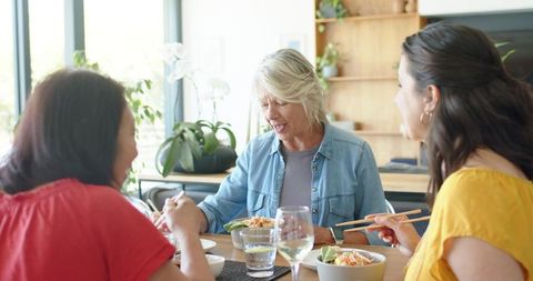 Diverse women sharing sunlit home meal with chopsticks, bowls and wine, casual friendship