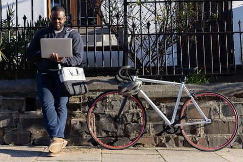 African american commuter working on laptop while leaning on stone wall beside bicycle