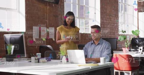 Coworkers collaborating over laptop in modern brick office showing social media reactions