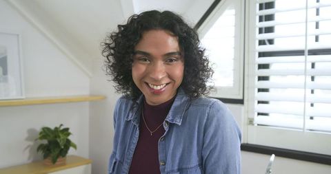 Non-binary adult smiling in bright loft wearing denim shirt and pendant, casual portrait