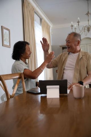 Senior asian couple high-fiving at dining table with tablet, cozy contemporary lifestyle