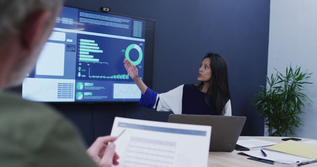 Businesswoman using large screen to present data analysis in a modern office environment. Colleague holding printed data in foreground. Can be used for business, corporate training, data presentation, teamwork, collaboration themes, and marketing materials.